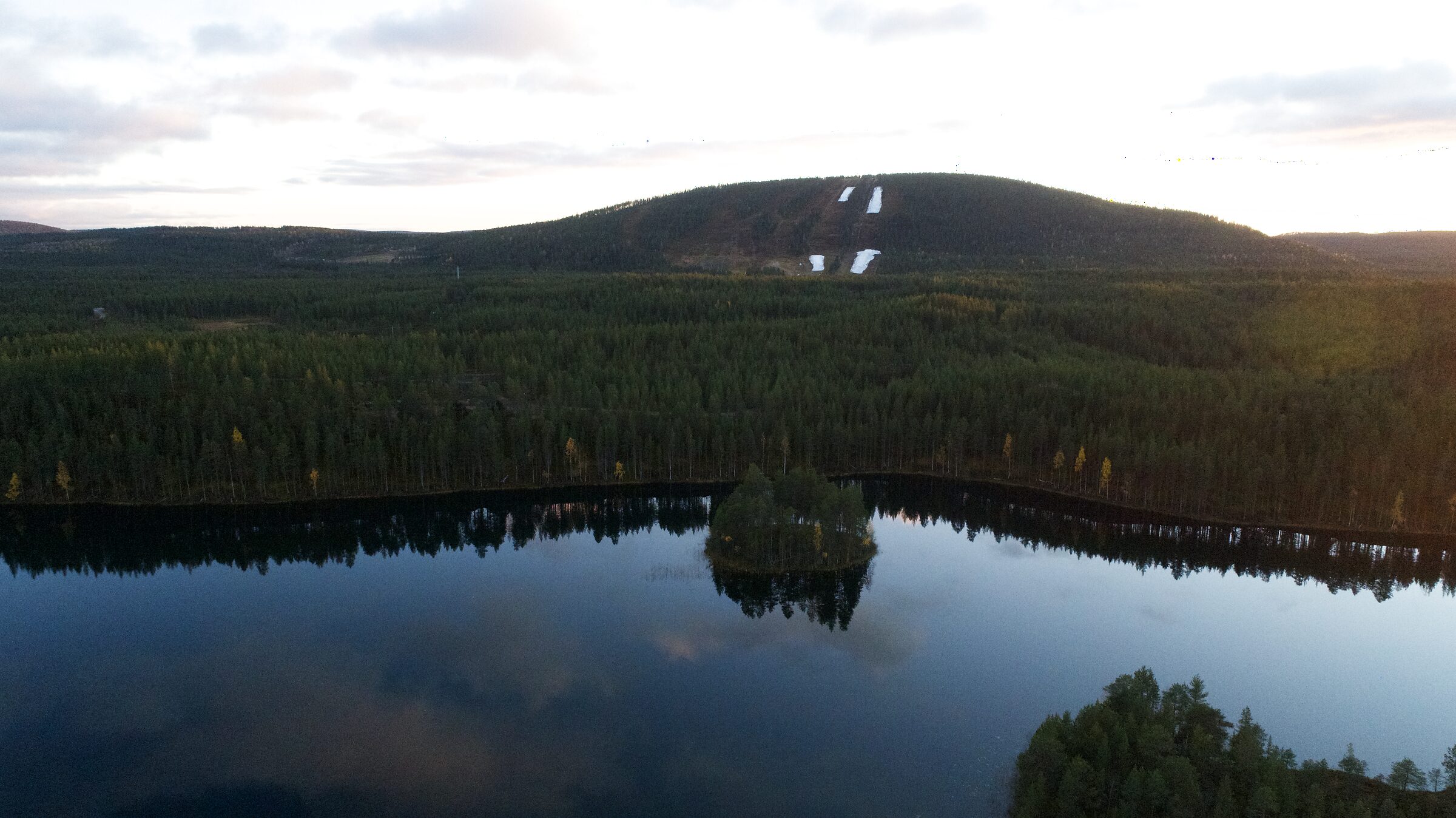 Luftaufnahme des Herajärvi mit seiner kleinen Insel, dahinter Kiefernwald und die Hänge des Suomutunturi. Die Lage von Hera Resort ist zwischen den Bäumen am gegenüberliegenden Ufer markiert.